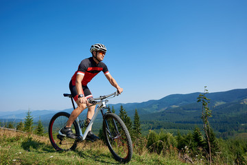 Young athletic tourist cyclist in helmet, sunglasses and full equipment riding bike down grassy hill on distant mountains and blue summer sky background. Active lifestyle and extreme sport concept.