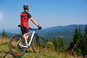 Obraz premium Back view of athletic professional bicyclist stopped on top of hill to enjoy beautiful view of distant Carpathian mountains on blue summer sky background. Active lifestyle and extreme sport concept.
