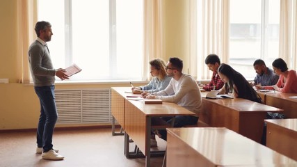 Male professor in casual clothes is talking to group of students sitting at tables in classroom and making notes. Large lecture hall with desks, chairs and windows is visible. - Powered by Adobe