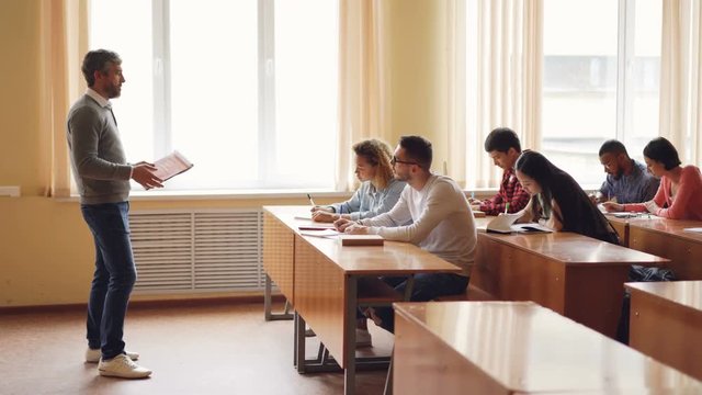 Male professor in casual clothes is talking to group of students sitting at tables in classroom and making notes. Large lecture hall with desks, chairs and windows is visible.