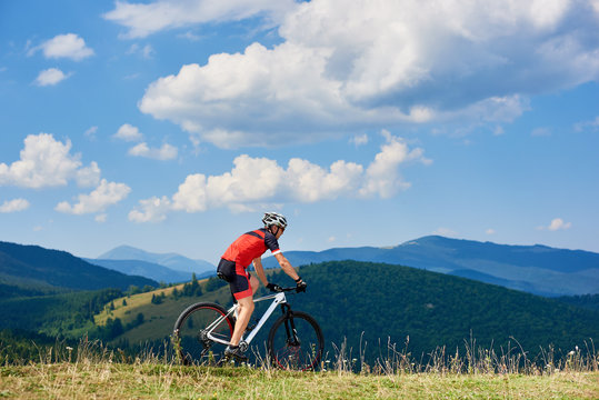 Professional sportsman cyclist in sportswear and helmet riding cross country bike in high grassy hill. Beautiful mountains view and cloudy sky on background. Active lifestyle and extreme sport concept