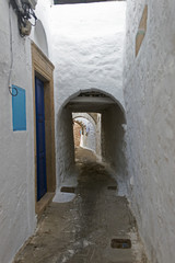 Fototapeta na wymiar A view of a narrow street with arch and wooden windows and doors with white wall stone architecture of the island Patmos, Greece