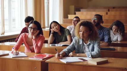 Tired and bored students are listening to teacher and making notes in notebooks sitting at desks in university. Education, boredom, young people and classroom concept.