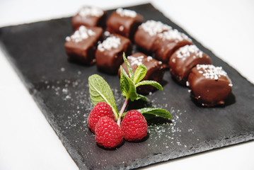 Dessert of red raspberries and chocolates, beautifully decorated on a graphite blackboard tray and sprinkled with coconut, on a white background.