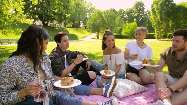 friendship, leisure and fast food concept - group of happy friends eating sandwiches or burgers and drinking non alcoholic drinks at picnic in summer park