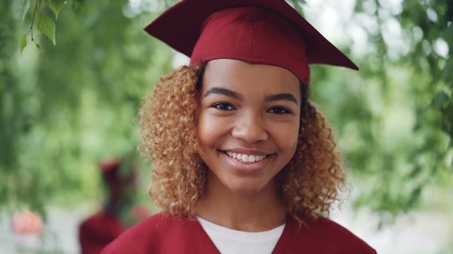 Slow motion portrait of pretty African American girl graduating student in red gown and mortarboard standing outdoors, smiling and looking at camera. Youth and education concept.