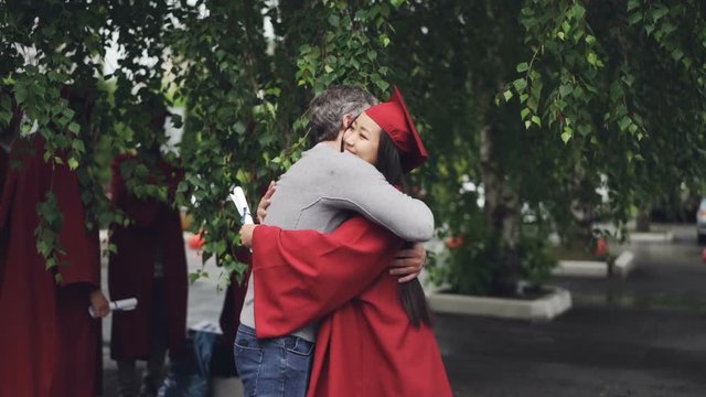 Loving father is congratulating his daughter on graduation day, people are hugging and laughing outdoors while other graduates are visible in background.