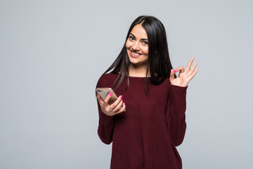 Attractive young girl winking and showing ok gesture while holding blank screen mobile phone over...