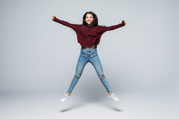 Fototapeta premium Pełnometrażowy portret ładnej, dziewczęcej kokietki, uroczej, nowoczesnej, stylowej dziewczyny w dżinsowym stroju, spodniach, skaczącej w powietrzu z uniesioną nogą, patrząc na kamery, na białym tle na szarym tle