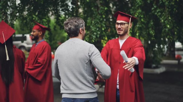 Smiling graduating student is shaking his father's hand and hugging him, young man in glasses is wearing hat and gown and holding diploma. Education and success concept.
