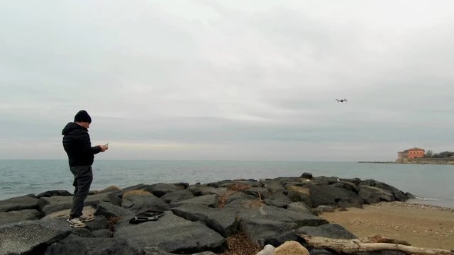 A man, in a winter cloudy day , on a rock beach with a seaview, drive a drone moving away over the sea