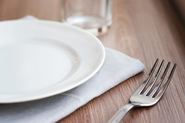 Empty white plate on a gray napkin with fork and glass on brown table. Table setting, preparation for meals.