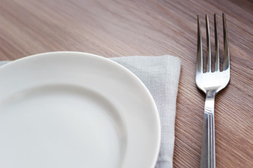 Empty white plate on a gray napkin with fork and glass on brown table. Table setting, preparation for meals.