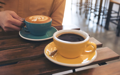 Closeup image of a woman holding a blue cup of hot coffee and another yellow cup on table in cafe
