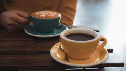 Closeup image of a woman holding a blue cup of hot coffee and another yellow cup on table in cafe