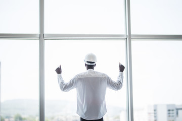 Young afro american man in protective hemlet standing in front of panoramic windows with reaised hands of victory and sucess