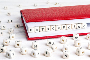 Word Outsourcing written in wooden blocks in red notebook on white wooden table.