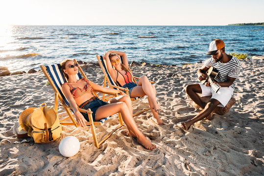 beautiful young women resting on beach chairs and african american man playing guitar