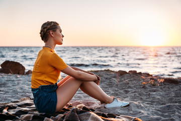 side view of pensive young woman sitting on plaid at beautiful sea coast at sunset