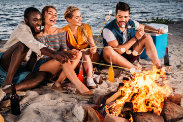 happy young multiethnic friends roasting marshmallows at bonfire on beach at sunset