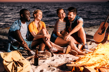 happy young multiethnic friends talking and spending time together on sandy beach at sunset