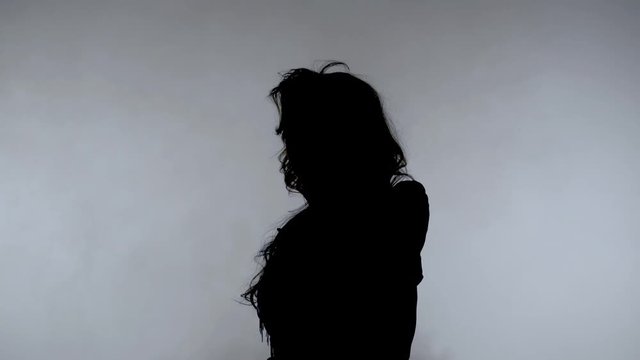 Young woman dark silhouette on smoke background. Beautiful woman with long hair rotating on gray fog background. Mysterious girl black silhouette. Young woman silhouette in scenic dress.