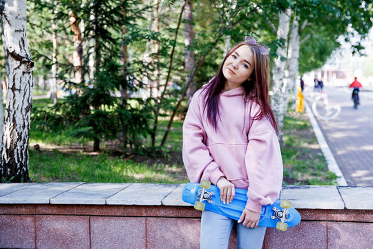 A young girl sits at the stairs in a pink sweatshot and holds a skateboard in her hands. Rest after a ride on a skateboard.