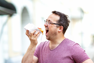 Closeup portrait of hungry man in glasses eating hot dog at outdoors background.