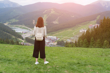 Fototapeta na wymiar Girl with a backpack standing on the hill and admiring the mountains. Town in the distance