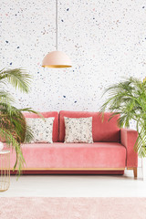Pastel lamp above pink settee with pillows in patterned living room interior with plants. Real photo