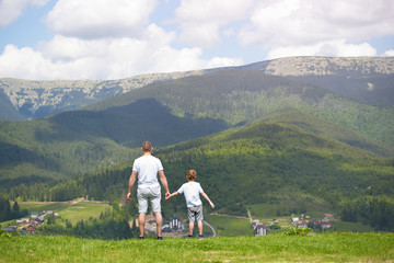 Father and young son standing on the lawn and admiring the mountains. Summer sunny day. Back view