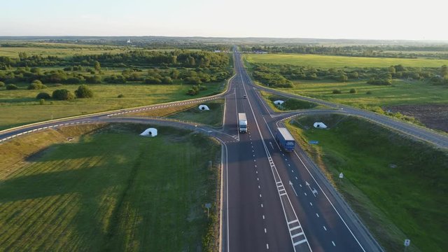 Aerial view. The cargo traffic on the suburban highway. Trucks carry various cargoes along roads. Heavy vehicles transport large pipes for pipelines.  Long evening shadows fall from cars on asphalt.