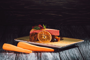 close up view of carrot cake with berry filling, mint, orange slice on plate near carrots on wooden table