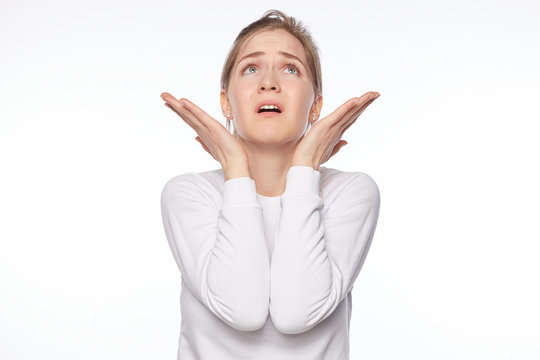 God please. Close up isolated portrait of young unhappy Caucasian female student dressed in white jumper holding palms up, looking up with worried expression, asking why and begging for happy end.