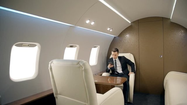 Young successful businessman is using smartphone while sitting in private plane, elegant business person in suit with tie is looking at device screen during business trip. Concept: entrepreneurship