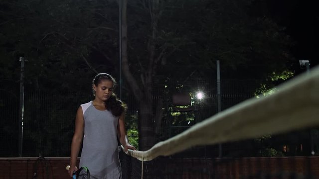The girl tennis player walks along the tennis net. Playing tennis at night.