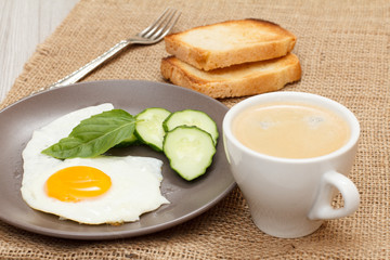 Plate with fried egg, fresh cut cucumber and leaf of basil, cup of black coffee, toasts and fork