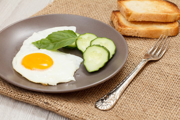 Plate with fried egg, fresh cut cucumber and leaf of basil, toasts and fork