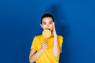 Beautiful surprised brunette girl holding lollipop on blue background