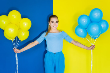 Smiling blonde woman holding blue and yellow balloons on blue and yellow background © LIGHTFIELD STUDIOS