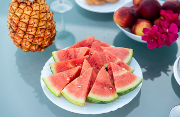 View of tasty breakfast on glass table, juice fruits, nectarine, watermelon, pineapple, water and bake. Healthy, colorful and exotic food in paradise. Summertime concept. Palm leaf on background.