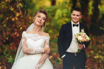 A thoughtful and romantic bride embraces herself in the autumn forest. in the background the groom...