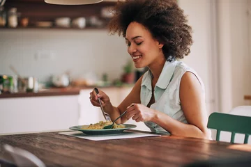 Gordijnen Beautiful mixed race woman eating pasta for dinner while sitting at kitchen table. © chika_milan