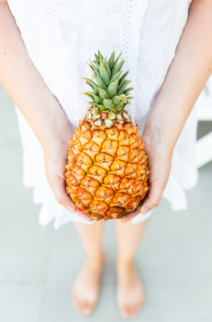 View of incognito female in white dress holding fresh and tasty pineapple in hands against wall background with palm leaf. Concept of fresh exotic fruits and raw summer food.