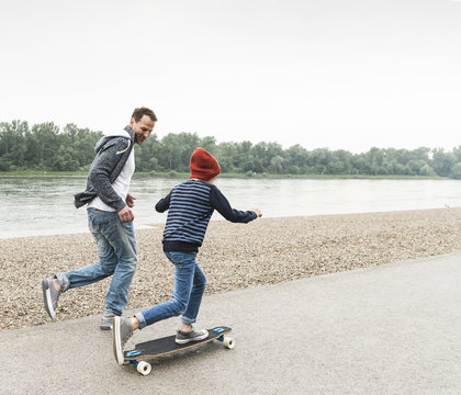 Happy father running next to son on skateboard at the riverside