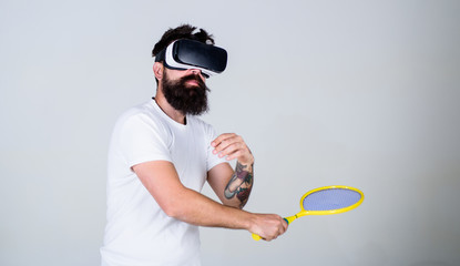Fototapeta na wymiar Man with beard in VR glasses beating pitch, grey background. Guy with VR glasses play tennis with racket. Virtual sport concept. Hipster on busy face use modern technology for sport games