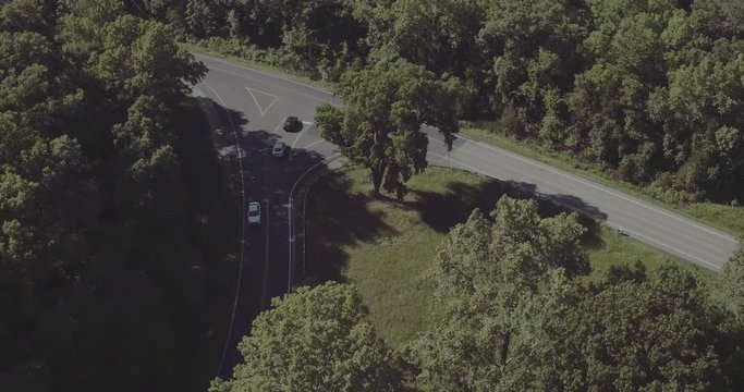 Drone footage of cars entering and leaving 3 way intersection in between tall trees while rotating around it.