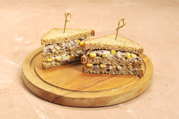 Two sandwiches of triangular shape on a wooden tray. Sandwiches are fastened with skewers. The filling of the sandwich consists of corn and tuna. Close-up. Light background.