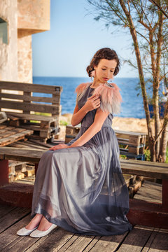 a young nymphet sits in a dress on a bench with the sea on the background and pulls the feathers on her dress