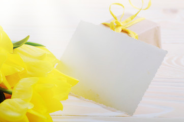 Yellow tulips near blank greeting card and gift box on natural wooden background with space for text